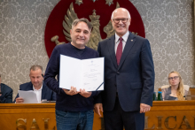 Dwie osoby stoją przed stołem prezydialnym. Jedna z nich trzyma otwarty dokument skierowany w stronę aparatu. W tle znajdują się uczestnicy obrad.