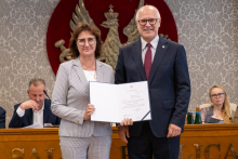 Dwie osoby pozują do zdjęcia z otwartym dokumentem. Za nimi widoczne są osoby siedzące przy stole oraz godło na ścianie.