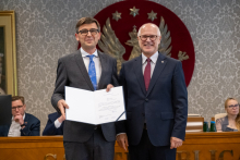 Dwie osoby stoją obok siebie. Jedna prezentuje otwartą teczkę z dokumentem. W tle znajdują się uczestnicy posiedzenia.