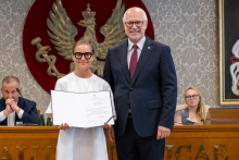 Dwie osoby pozują do zdjęcia przed godłem. Jedna z nich trzyma otwarty dokument. W tle widoczne są osoby siedzące przy stole prezydialnym.