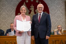 Dwie osoby pozują do zdjęcia. Jedna z nich trzyma otwartą teczkę z dokumentem skierowanym do aparatu. W tle widoczne są osoby uczestniczące w obradach.