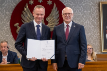 Dwie osoby stoją przed stołem prezydialnym. Jedna z nich trzyma otwartą teczkę z dokumentem skierowanym do aparatu. W tle widoczne są osoby uczestniczące w posiedzeniu oraz godło na ścianie.