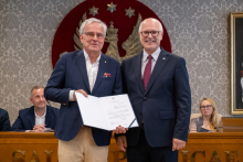 Dwie osoby pozują do zdjęcia i prezentują otwarty dokument. W tle znajdują się uczestnicy obrad siedzący przy stole prezydialnym.