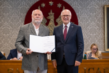 Dwie osoby stoją obok siebie. Jedna z nich trzyma otwartą teczkę z dokumentem. W tle widoczne są osoby uczestniczące w posiedzeniu oraz godło.