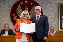 Dwie osoby stoją przed godłem i prezentują otwarty dokument. W tle znajdują się osoby siedzące przy stole prezydialnym.