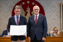 Dwie osoby pozują do zdjęcia. Jedna z nich trzyma otwartą teczkę z dokumentem skierowanym do aparatu. W tle widoczne są uczestnicy obrad.