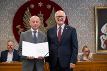 Dwie osoby stoją obok siebie. Jedna prezentuje otwarty dokument. W tle znajdują się osoby siedzące przy stole prezydialnym.