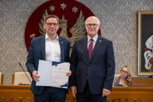 Dwie osoby pozują do zdjęcia. Jedna z nich trzyma otwartą teczkę z dokumentem. W tle widoczne są uczestnicy posiedzenia oraz elementy wyposażenia sali.