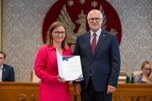 Dwie osoby stoją przed stołem prezydialnym. Jedna z nich prezentuje dokument w otwartej teczce. W tle znajdują się uczestnicy obrad.