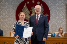 Dwie osoby pozują do zdjęcia. Jedna trzyma otwartą teczkę z dokumentem. W tle widoczne są osoby siedzące przy stole prezydialnym.