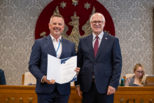 Dwie osoby stoją obok siebie i prezentują otwarty dokument. W tle znajdują się uczestnicy posiedzenia oraz godło na ścianie.