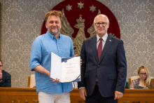 Dwie osoby pozują do zdjęcia z dokumentem w otwartej teczce. W tle widoczne są osoby uczestniczące w obradach.