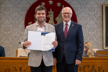 Dwie osoby stoją przed stołem prezydialnym. Jedna z nich trzyma otwartą teczkę z dokumentem skierowanym do aparatu. W tle znajdują się uczestnicy posiedzenia.