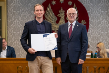 Dwie osoby pozują do zdjęcia i prezentują dokument w otwartej teczce. W tle widoczne są osoby siedzące przy stole prezydialnym oraz godło.