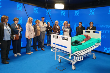 Grupa kilkunastu osób w pomieszczeniu do symulacji medycznych. Osoby stoją koło łóżka szpitalnego, na którym leży fantom do ćwiczeń.