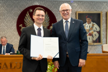 Dwie osoby stoją obok siebie i prezentują otwarty dokument. W tle widoczne są stół prezydialny, osoby uczestniczące w posiedzeniu oraz godło na ścianie.