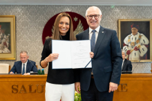Dwie osoby stoją obok siebie i trzymają otwarty dokument skierowany w stronę aparatu. W tle znajdują się uczestnicy posiedzenia, stół prezydialny oraz godło umieszczone na ścianie sali.