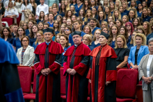 A woman and two men in red robes are standing among the ceremony attendees. In the background, male and female students are seated in navy blue medical uniforms.