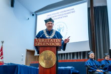 A man in a blue academic gown stands at the podium and delivers a speech during the cap-and-gown ceremony for the nursing and midwifery programs.