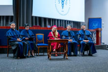 A woman and four men in togas are seated on stage during the ceremony. The woman is wearing a red toga; the others are wearing blue ones.