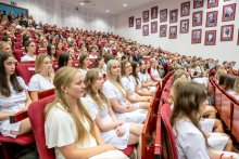 Graduates in white medical scrubs are seated in rows in the auditorium during the ceremony. The other attendees are in the background.