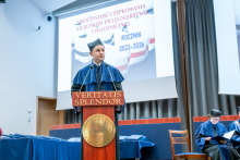 Mężczyzna w niebieskiej todze przemawia przy mównicy podczas uroczystości czepkowania. Na ekranie za nim wyświetlona jest nazwa wydarzenia.