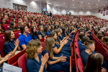 Absolwentki i absolwenci w granatowych strojach medycznych siedzą w auli i biją brawo.