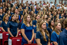 Graduates in navy blue medical uniforms stand between the rows of seats during the official part of the ceremony.
