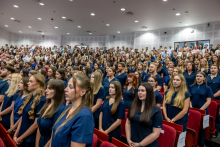 Grupa absolwentek i absolwentów w granatowych strojach medycznych stoi w auli. W tle znajdują się pozostali uczestnicy wydarzenia.