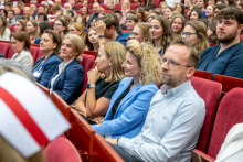 Women and men are seated among the audience during the ceremony. In the foreground is a woman wearing a light blue blazer.