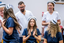 Absolwentka w granatowym stroju medycznym i czepku pielęgniarskim siedzi i bije brawo. W tle stoją kobiety i mężczyzna w białych strojach medycznych.