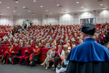 A view of the auditorium during the cap-and-gown ceremony. In the foreground stands a man in a blue academic gown, and the audience fills nearly every seat in the hall.