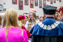 A young woman in a white medical uniform and a midwife's cap is receiving congratulations from a woman in a red gown. In the background, the ceremony's attendees are standing.