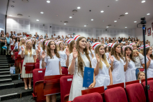 Grupa kobiet w białych strojach medycznych i czepkach położnych stoi w rzędach auli z uniesionymi prawymi dłońmi podczas ślubowania. Jedna z kobiet trzyma w ręku niebieską