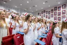 Women in white medical scrubs and midwife caps stand with their right hands raised as they take the oath. Blue folders rest on the backs of the chairs.