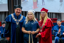 A man and a woman in robes are standing on either side of a young woman wearing a navy blue medical uniform and a nurse's cap. The woman is holding a beige cloth bag.