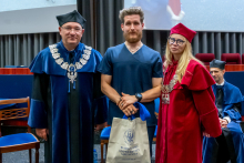Mężczyzna w niebieskiej todze i kobieta w czerwonej todze pozują do zdjęcia z młodym mężczyzną w granatowym stroju medycznym. Mężczyzna trzyma w rękach papierową torbę z logo Warszawskiego Uniwersytetu Medycznego.