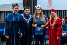 Two men and a woman in robes are posing for a photo with a young woman wearing a navy blue medical uniform and a nurse's cap. The woman is holding a blue briefcase and a navy blue bag.