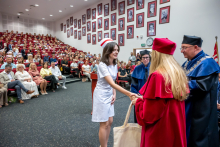 A young woman in a white medical uniform and a midwife's cap is receiving congratulations from a woman in a red gown. Standing next to them are a woman and a man in blue gowns. In the background, an audience gathered in the auditorium can be seen.