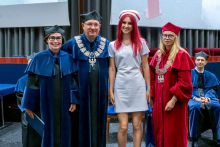 A woman and a man in blue gowns and a woman in a red gown are posing for a photo with a young woman wearing a white medical uniform and a midwife's cap.
