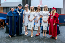 A woman and a man in blue gowns and a woman in a red gown are standing with four young women in white medical uniforms and midwife caps. The women are holding white bags.