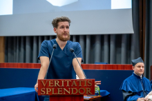 A young man in a navy blue medical uniform stands at the podium and speaks during the ceremony. In the background, a man in a blue robe is sitting.