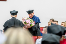 Mężczyzna w niebieskiej todze odbiera bukiet kwiatów od osoby stojącej tyłem do obiektywu. Wokół siedzą uczestnicy uroczystości.