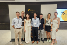 A group of seven people wearing conference badges pose for a photograph in front of a display celebrating the 75th anniversary of the Lindau Nobel Laureate Meetings. The display behind them features the slogan “Science Transcending Borders.”