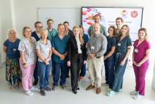 A group of medical staff and event participants pose for a group photograph in a conference room. A presentation screen about robotic surgery in children is visible in the background.