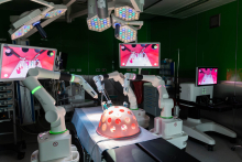 A robotic surgery workstation equipped with four robotic arms and monitors displaying an internal surgical view. Operating lights are suspended above the workstation.