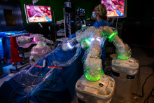 A person operates a robotic surgery console during a procedure. Robotic arms are positioned around the patient, and surgical images are displayed on the monitors.