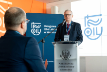 Mężczyzna, Rzecznik Praw Lekarzy Naczelnej Izby Lekarskiej, stoi przy mównicy z logo Warszawskiego Uniwersytetu Medycznego i przemawia do uczestników konferencji organizowanej we współpracy z Okręgową Izbą Lekarską w Warszawie.
