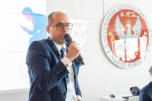 Mężczyzna z mikrofonem, dr Artur Drobniak, stoi przed ekranem multimedialnym. Na ekranie widoczne są logotypy Warszawskiego Uniwersytetu Medycznego i Okręgowej Izby Lekarskiej w Warszawie.