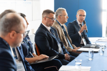 Pięć osób siedzi przy stole panelowym. Jedna z osób zabiera głos podczas dyskusji prowadzonej w trakcie konferencji.